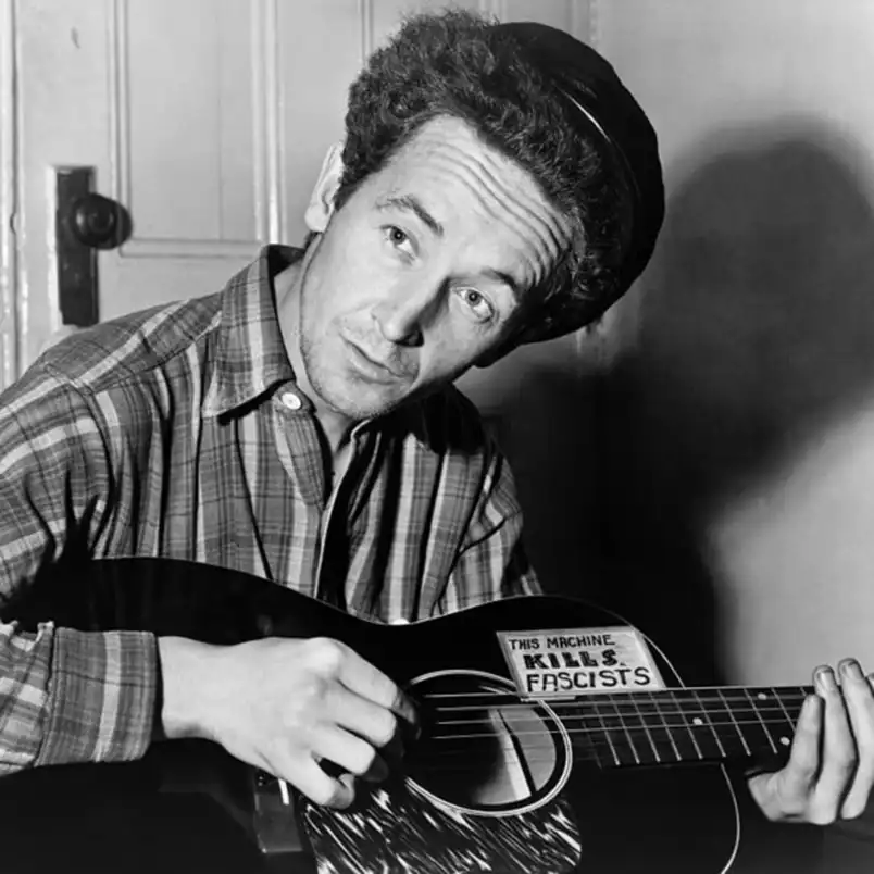 Woody Guthrie com um violão com um adesivo escrito "Esta máquina mata fascistas"